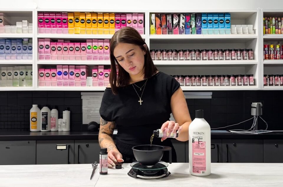 Hairstylist mixing professional hair color in a salon environment with products on display behind her